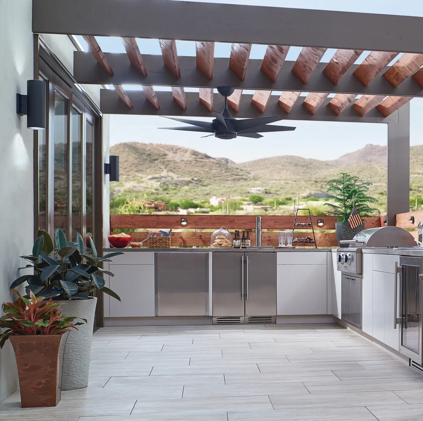 Cocina exterior bajo pérgola de madera con ventilador de techo negro y vista a las montañas del desierto, de día