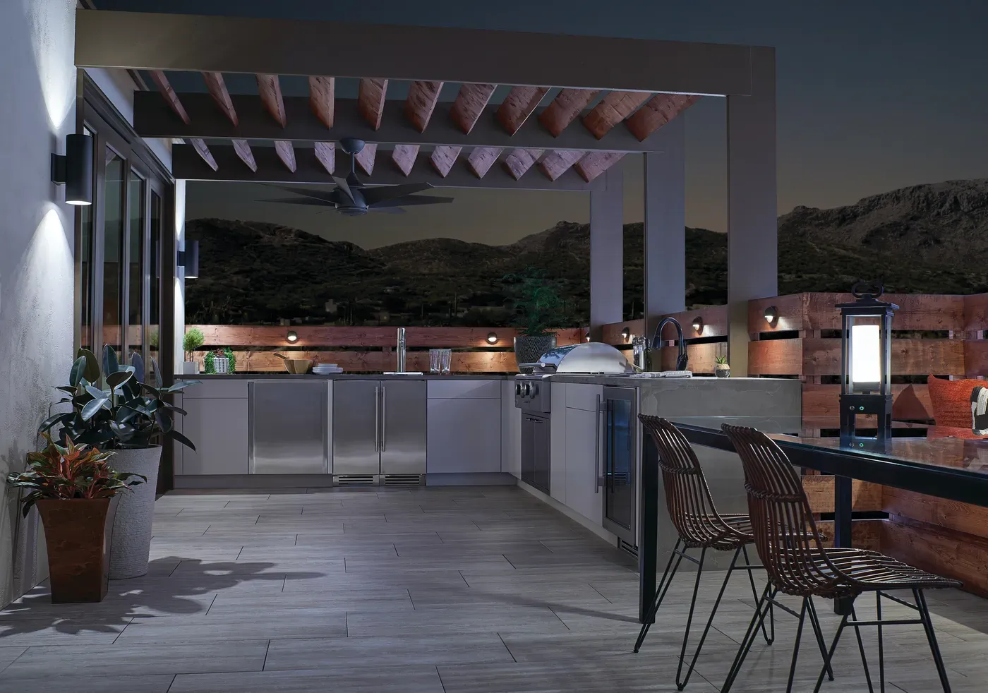 La misma cocina exterior al anochecer, con el ventilador de techo negro y la iluminación LED encendida bajo la pérgola